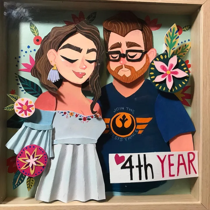 Layered paper shadow box portrait of a couple, a woman in a blue ruffled dress and a bearded man, with a 4th Year sign.