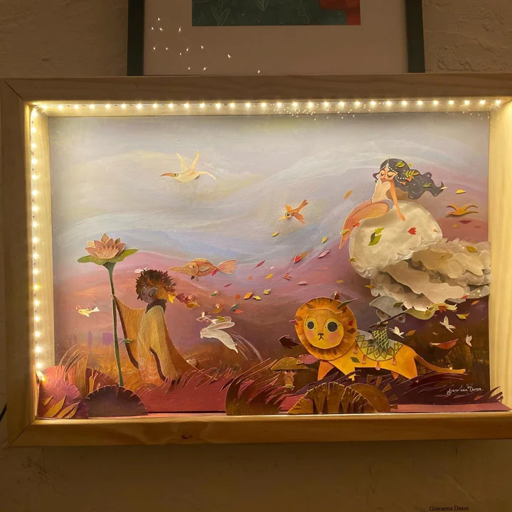 Lit paper-cut shadow box of a robed girl, a lion, birds, and a mermaid on a jellyfish under a pink sky, in a wood frame.