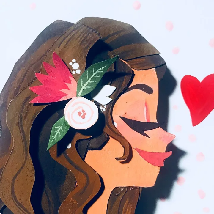 Layered paper-cut profile of a woman with flowers in her brown hair, blowing a kiss toward a red heart.