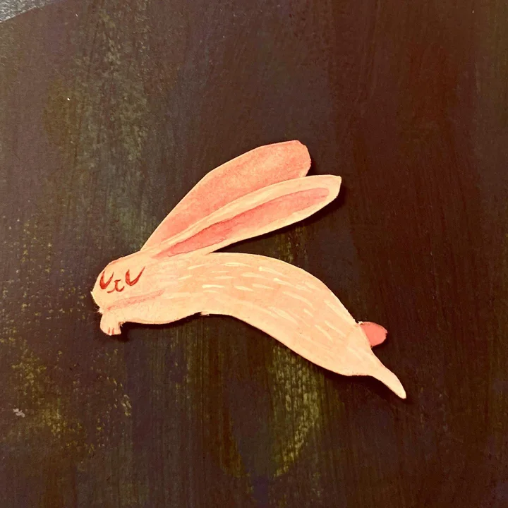 Painted paper cut-out of a leaping peach-colored rabbit with long ears, on a dark wood surface.