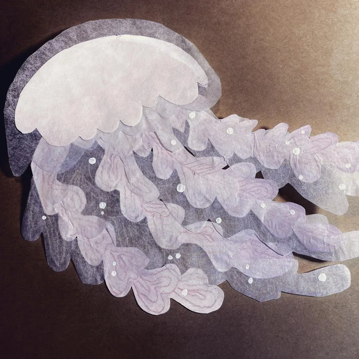 Tissue-paper cut-out of a jellyfish in white and lavender with ruffled tentacles on a dark background.