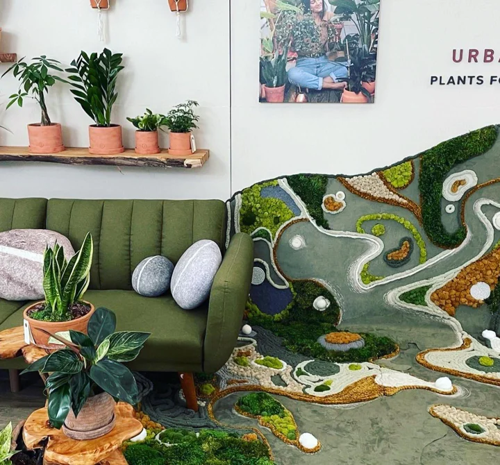 Photo of a moss, stone, and pebble landscape installation flowing from a plant-shop wall onto the floor beside a green sofa.