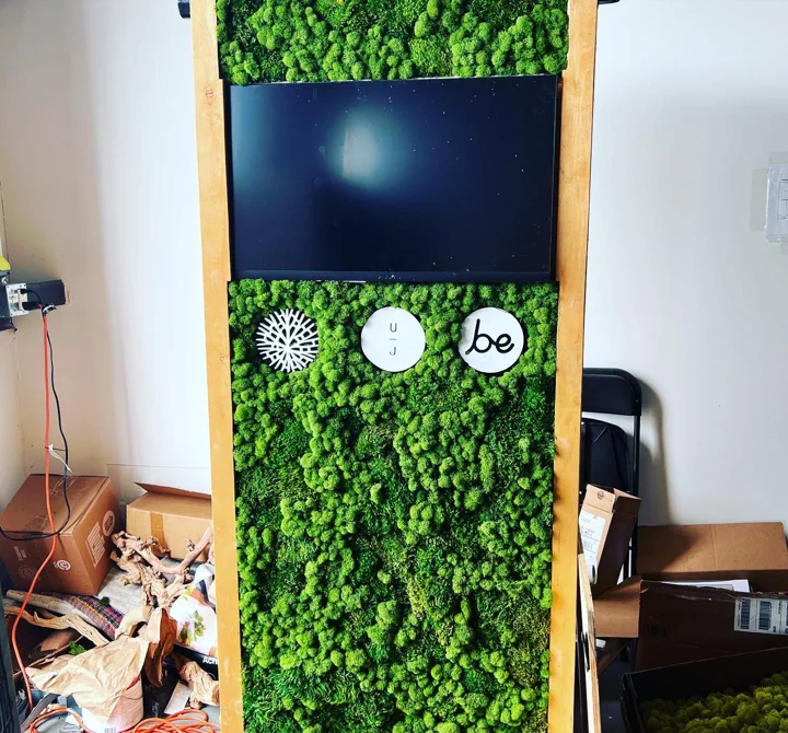 Photo of a tall wood-framed moss wall with a mounted screen and three round logo plaques, in a cluttered work room.