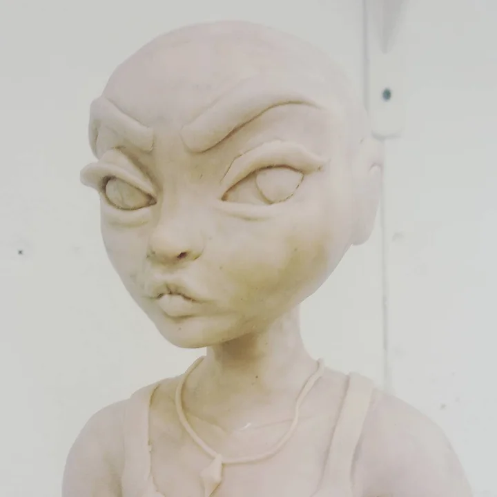 Photo of an unpainted clay bust of a bald, wide-eyed girl with a furrowed brow and pursed lips, wearing a tank top.