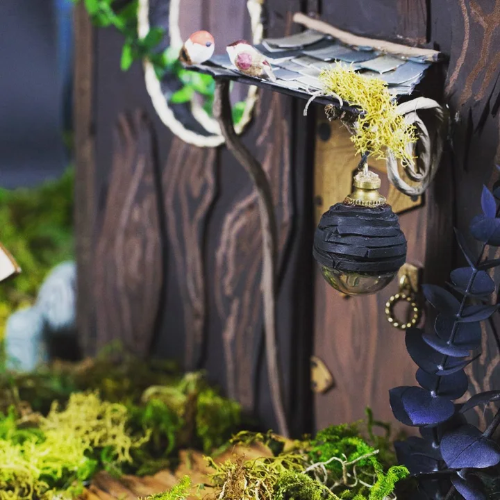 Photo of a handmade miniature fairy house door with a hanging lantern, a tiny bird on a shingled awning, and moss.
