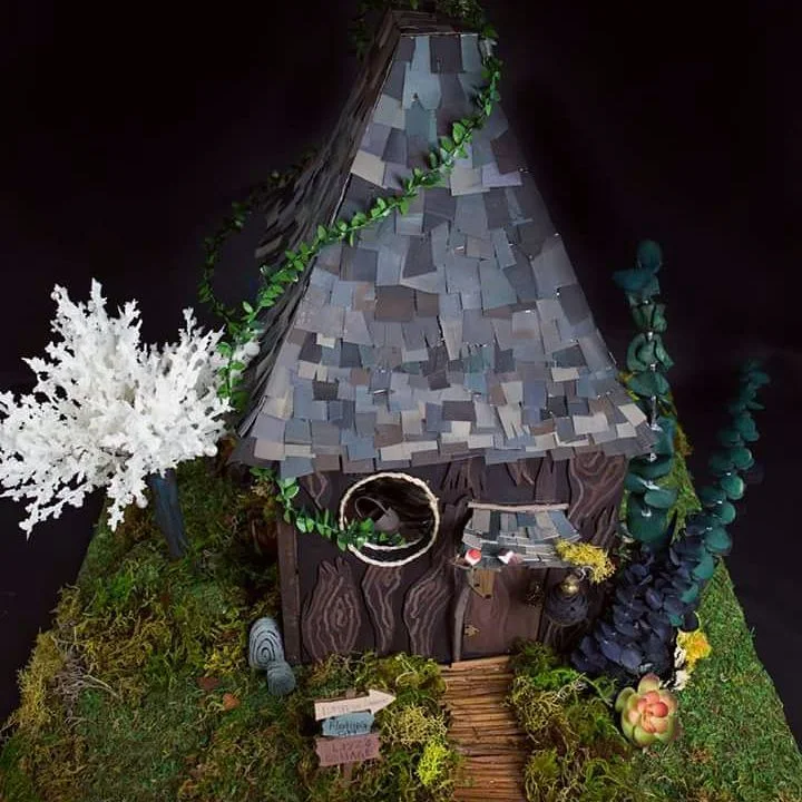 Photo of a handmade miniature fairy cottage with a shingled cone roof, a round window, climbing vines, moss, and a signpost.