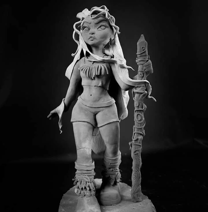 Black-and-white photo of a clay figure of a young warrior woman with wild hair, a fringed top, and a carved staff.